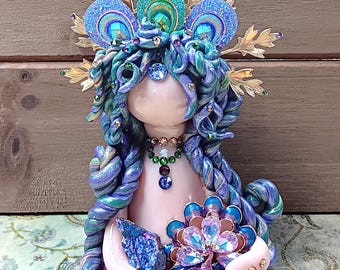 May include: A handcrafted figurine of a seated figure with a pale pink body and blue and purple hair. The figure wears a jeweled crown and necklace. It holds a blue crystal and a jeweled peacock-like flower. The figurine is on a patterned surface.