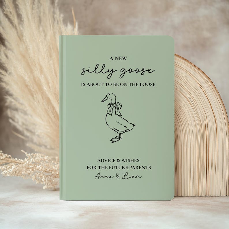 Op de afbeelding: Een saliegroen notitieboek met de tekst "A new silly goose is about to be on the loose." Een lijntekening van een gans staat onder de tekst. Het notitieboek is voor advies en wensen voor toekomstige ouders, Anna & Liam.