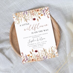 以下が含まれることがあります： 花柄のベビーシャワー招待状。「A little wildflower is on the way」と書かれています。招待状には、お祝いされる方の名前、日付、時間、場所が記載されています。