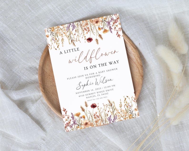 以下が含まれることがあります： 花柄のベビーシャワー招待状。「A little wildflower is on the way」と書かれています。招待状には、お祝いされる方の名前、日付、時間、場所が記載されています。