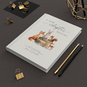 May include: A white journal with the text "A New Chapter Begins" and a watercolor illustration of woodland animals. The journal includes the text "Advice & Wishes for the Future Parents". Gold and black pencils and binder clips are also visible.
