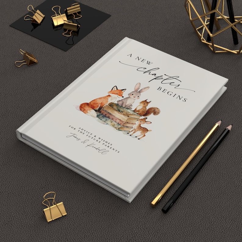 May include: A white journal with the text "A New Chapter Begins" and a watercolor illustration of woodland animals. The journal includes the text "Advice & Wishes for the Future Parents". Gold and black pencils and binder clips are also visible.