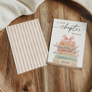 May include: A wooden tray holds a card with the text "A New Chapter Begins" above a stack of books with a pink bow. A second card with vertical beige stripes is also present.