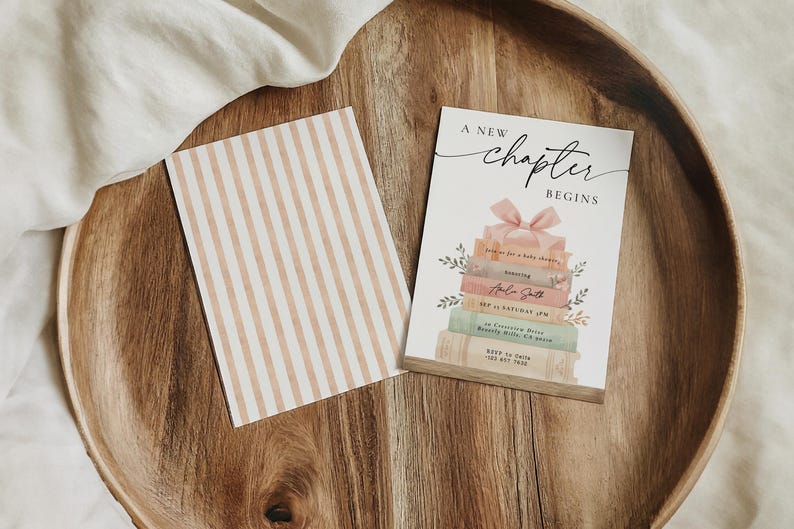 May include: A wooden tray holds a card with the text "A New Chapter Begins" above a stack of books with a pink bow. A second card with vertical beige stripes is also present.