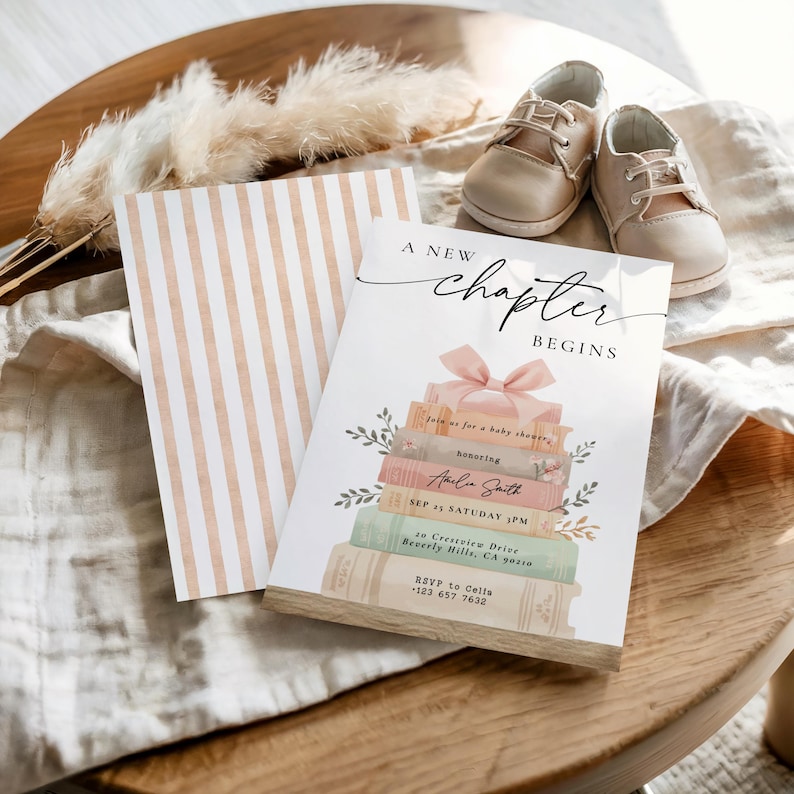 May include: A baby shower invitation with the text "A New Chapter Begins" and a stack of books with a pink bow. The invitation is accompanied by a striped card and a pair of beige baby shoes, all arranged on a wooden surface.