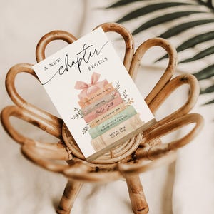 May include: A baby shower invitation card with the text "A new chapter begins" and a stack of books design. The card is decorated with a pink bow and floral accents, resting on a woven wooden chair.
