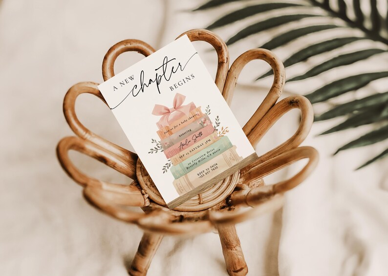 May include: A baby shower invitation card with the text "A new chapter begins" and a stack of books design. The card is decorated with a pink bow and floral accents, resting on a woven wooden chair.