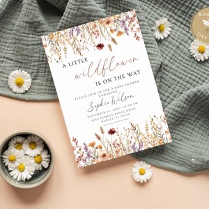 以下が含まれることがあります： ベビーシャワーの招待状。テキストは「A Little Wildflower Is On The Way」とあり、Sophie Wilsonのベビーシャワーの詳細が記載されています。招待状は、紫、黄色、茶色の水彩画の花で囲まれています。