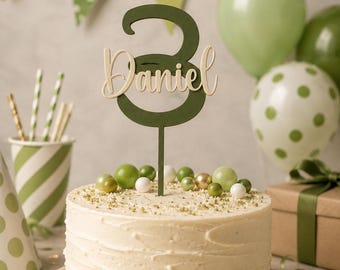 Pode incluir: Um bolo redondo com cobertura branca, decorado com um topo de bolo em forma de número três verde-escuro com o nome Daniel em letras cursivas claras. O bolo é enfeitado com pequenas esferas verdes e brancas e granulado.
