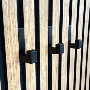 May include: Three black metal hooks mounted on a wooden panel with vertical wood grain. The hooks are square and project from the panel. The panel has alternating light wood and black stripes.