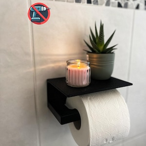 Può includere: Un portarotolo in metallo nero opaco con ripiano piatto integrato. Il ripiano sostiene una piccola pianta grassa verde in un vaso grigio e una candela rosa accesa in un barattolo di vetro. È visibile un'icona che indica senza trapano.