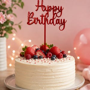 Op de afbeelding: Een rode acryl taarttopper met de tekst Happy Birthday in een cursief lettertype. De topper staat bovenop een ronde taart met wit glazuur, versierd met verse aardbeien, bosbessen en kleine witte en roze suikerkorrels als decoratie.