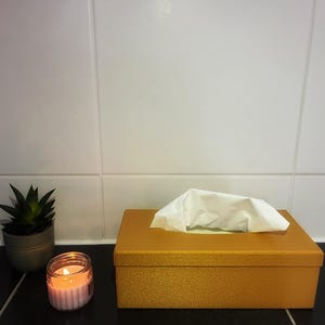 Könnte beinhalten: Eine goldene Taschentuchbox mit weißen Taschentüchern, eine kleine Topf-Sukkulente und eine brennende Kerze in einem Glas. Die Gegenstände stehen auf einer schwarzen Oberfläche vor einer weißen gefliesten Wand.