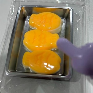 May include: Three fish-shaped, orange-yellow candies in a small, rectangular metal tray, inside a clear plastic package. The candies have a glossy appearance. A purple object is partially visible in the frame.