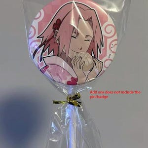 Op de afbeelding: Een ronde, roze badge met een manga-achtige illustratie van een personage met roze haar, dat eten vasthoudt. De badge is verpakt in doorzichtig cellofaan met een gouden lint en een witte stok.