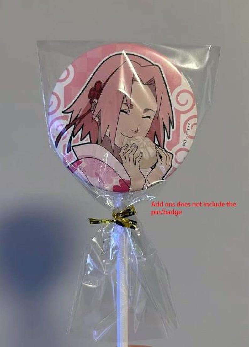 Op de afbeelding: Een ronde, roze badge met een manga-achtige illustratie van een personage met roze haar, dat eten vasthoudt. De badge is verpakt in doorzichtig cellofaan met een gouden lint en een witte stok.