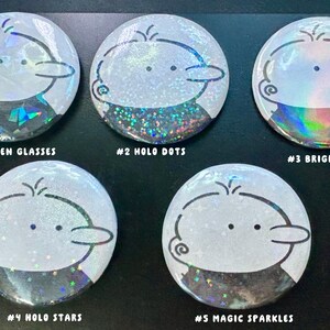 Op de afbeelding: Vijf ronde, witte buttons met een cartoonkarakterontwerp. Elke button heeft een ander holografisch effect, met tekstlabels: "#1 BROKEN GLASSES", "#2 HOLO DOTS", "#3 BRIGHT FILM", "#4 HOLO STARS" en "#5 MAGIC SPARKLES".