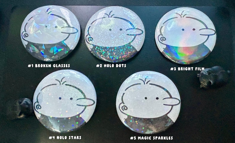 Op de afbeelding: Vijf ronde, witte buttons met een cartoonkarakterontwerp. Elke button heeft een ander holografisch effect, met tekstlabels: "#1 BROKEN GLASSES", "#2 HOLO DOTS", "#3 BRIGHT FILM", "#4 HOLO STARS" en "#5 MAGIC SPARKLES".