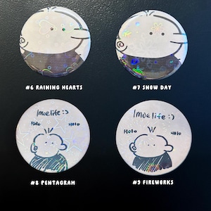 Op de afbeelding: Vier ronde, witte knopen met cartoon illustraties en tekst. Twee knopen hebben een ontwerp van een gezicht met de tekst "#6 RAINING HEARTS" en "#7 SNOW DAY". De andere twee knopen hebben een gezichtsontwerp met de tekst "#8 PENTAGRAM" en "#9 FIREWORKS".