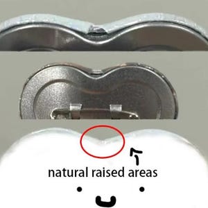 Op de afbeelding: Close-up van drie hartvormige buttonspins. De bovenste knop is zilverkleurig, de middelste knop is metallic en de onderste knop is wit met de tekst "natural raised areas" en een smiley.