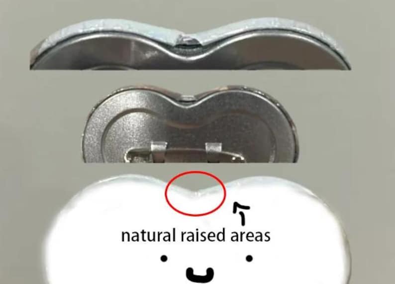Op de afbeelding: Close-up van drie hartvormige buttonspins. De bovenste knop is zilverkleurig, de middelste knop is metallic en de onderste knop is wit met de tekst "natural raised areas" en een smiley.