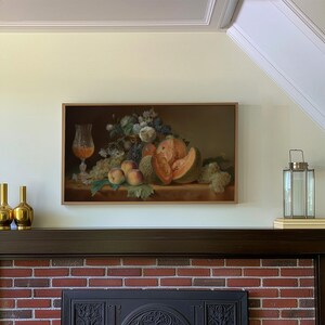 May include: A rectangular framed wall art print featuring a still life painting of a glass of amber liquid, white flowers, green grapes, peaches, and a sliced cantaloupe melon arranged on a surface against a dark, moody background.