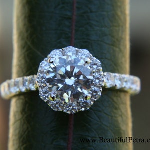 Op de afbeelding: Een zilveren verlovingsring met een grote ronde diamant omgeven door een halo van kleinere diamanten. De ring is gezet op een band met kleinere diamanten.
