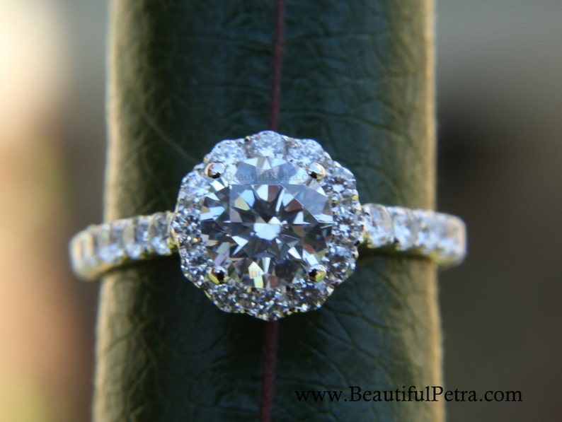 Op de afbeelding: Een zilveren verlovingsring met een grote ronde diamant omgeven door een halo van kleinere diamanten. De ring is gezet op een band met kleinere diamanten.