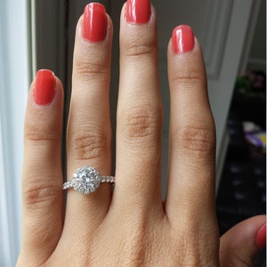 Op de afbeelding: Een close-up van een hand met rode nagellak die een zilveren ring draagt met een grote, ronde, heldere diamant omringd door kleinere diamanten.