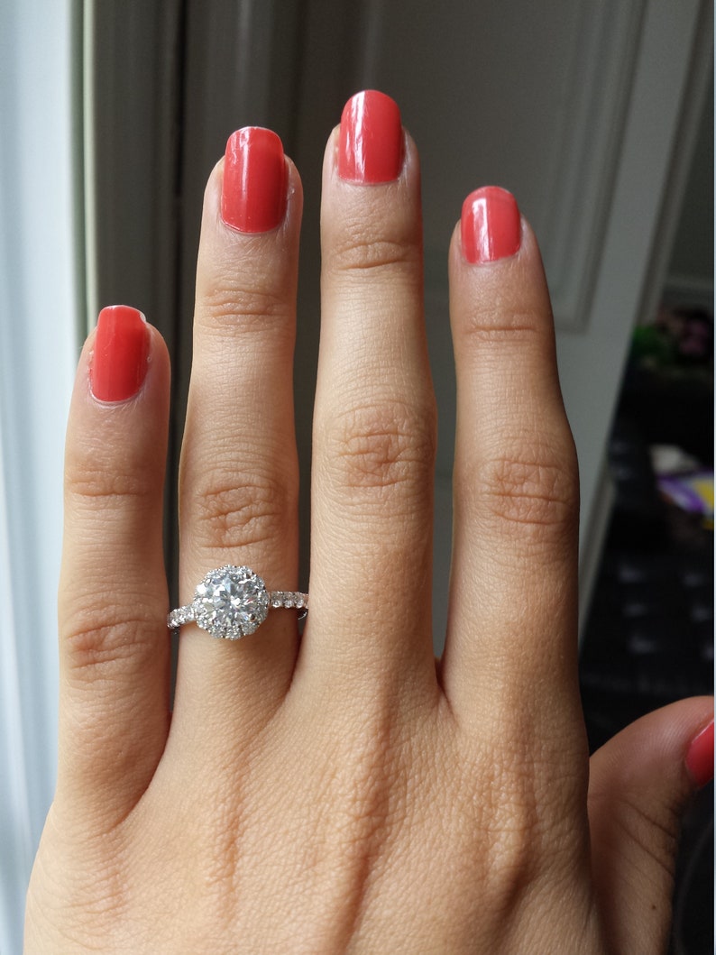 Op de afbeelding: Een close-up van een hand met rode nagellak die een zilveren ring draagt met een grote, ronde, heldere diamant omringd door kleinere diamanten.