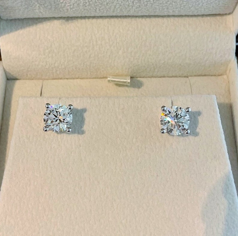 K&ouml;nnte beinhalten: Ein Paar runde, klare Diamant-Ohrstecker in einer wei&szlig;en Schachtel.