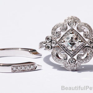 May include: A silver engagement ring with a square-shaped center stone and intricate filigree detailing. The ring is set with small diamonds and has a matching wedding band.