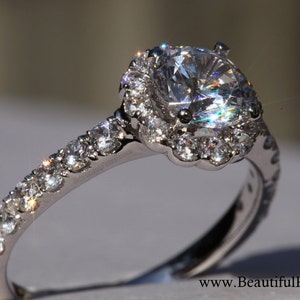 Op de afbeelding: Een zilveren verlovingsring met een grote, ronde, heldere diamant in het midden, omgeven door een halo van kleinere diamanten. De band is ook bezet met kleinere diamanten.