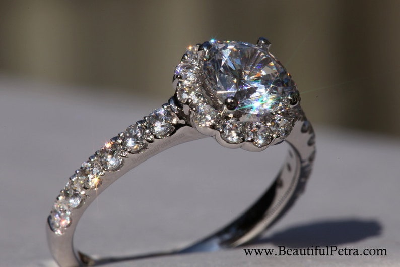 Op de afbeelding: Een zilveren verlovingsring met een grote, ronde, heldere diamant in het midden, omgeven door een halo van kleinere diamanten. De band is ook bezet met kleinere diamanten.