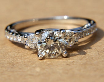 May include: A silver engagement ring with a large round center diamond and two smaller diamonds on either side. The ring is set with small diamonds around the band.