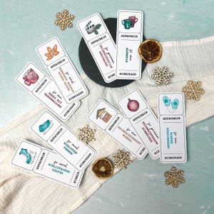 Könnte beinhalten: Eine Sammlung weißer Gutscheinkarten mit verschiedenen winterlichen Illustrationen, darunter Tassen, eine Filmrolle und Weihnachtsschmuck. Die Karten sind auf einem weißen Tuch mit Holzschneeflocken und getrockneten Orangenscheiben angeordnet.