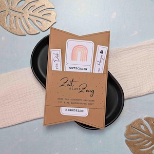 Puede incluir: Un tarjetero marrón con un arcoíris y la palabra "GUTSCHEIN". El tarjetero tiene el texto "Zeit statt Zeug" y "DENN DAS SCHÖNSTE GESCHENK IST EINE ZAUBERHAFTE ZEIT". El tarjetero está sobre una placa ovalada negra.