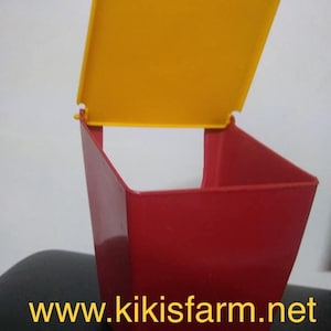 May include: Red plastic chicken feeder with a yellow lid. The feeder has a square base and a sloped top. The lid is hinged and can be opened to fill the feeder with food. The feeder is labeled with the website "www.kikisfarm.net".