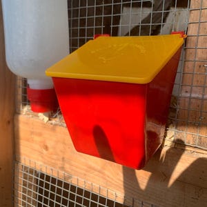May include: Red and yellow plastic chicken feeder with a hinged lid. The feeder has a white plastic bird silhouette on the lid with the text "Chickens & Roosters".