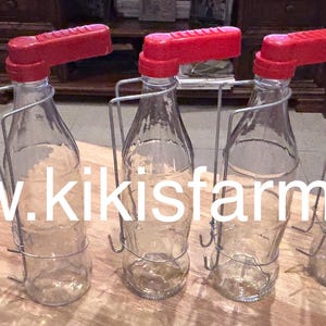 Może przedstawiać: Pięć przezroczystych szklanych butelek z czerwonymi wylewkami i metalowymi uchwytami drucianymi. Butelki są ułożone na drewnianej powierzchni. Adres strony internetowej www.kikisfarm.net jest wyświetlany białym tekstem na obrazie.