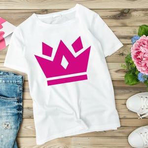 May include: White t-shirt with a pink crown graphic. The crown is made up of five points and has a white outline.