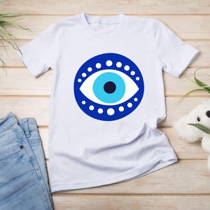 May include: White t-shirt with a blue evil eye design. The eye is surrounded by a circle of white dots.