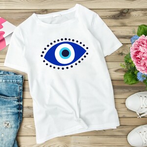May include: White t-shirt with a blue and white evil eye design. The evil eye is a symbol of protection against negative energy.