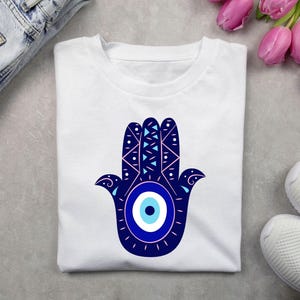 May include: White t-shirt with a blue and white hamsa hand design. The hamsa hand has a blue evil eye in the center.