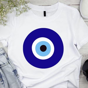 May include: White t-shirt with a blue evil eye design. The eye is surrounded by a white circle and a blue circle.