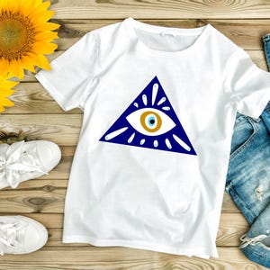 May include: White t-shirt with a blue triangle and a white eye with a blue pupil and a gold iris. The eye is surrounded by white rays.