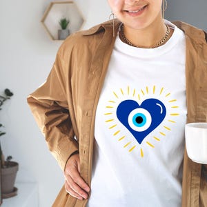 May include: White t-shirt with a blue heart featuring a blue and white evil eye design. The heart is surrounded by yellow rays.