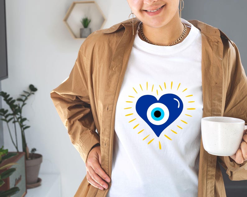May include: White t-shirt with a blue heart featuring a blue and white evil eye design. The heart is surrounded by yellow rays.