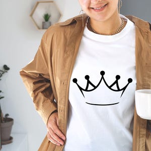May include: A white t-shirt with a black crown graphic. The shirt is being worn by a person with a brown jacket.