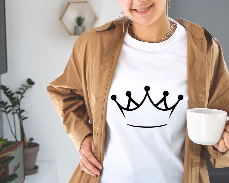 May include: A white t-shirt with a black crown graphic. The shirt is being worn by a person with a brown jacket.